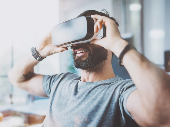 Bearded young man wearing virtual reality goggles in modern coworking studio. Smartphone using with VR headset in office. Horizontal, blurred.