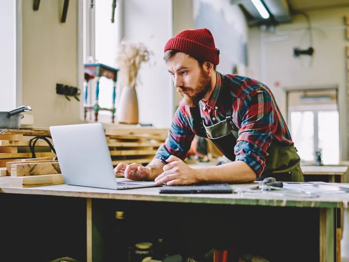 Young hipster guy watching training tutorial on laptop computer learning at carpentry courses, skilled male artisan using netbook for searching information during woodworks and making plannings.